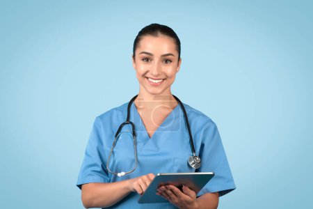 Photo for Professional woman nurse in blue scrubs, smiling while using digital tablet, exuding expertise and care in modern healthcare setting, with tranquil light blue background - Royalty Free Image