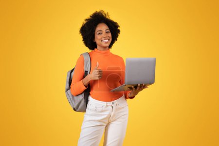 Photo for Cheerful african american young woman gives a thumbs-up while holding a laptop, wearing an orange turtleneck and white pants, with a gray backpack on a bright yellow background - Royalty Free Image