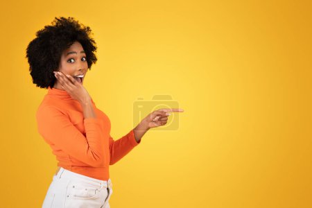 Téléchargez les photos : Surprise et ludique jeune femme afro-américaine gesticulant sur le côté d'une main, exprimant son excitation, portant un col roulé orange et un pantalon blanc, sur fond jaune - en image libre de droit