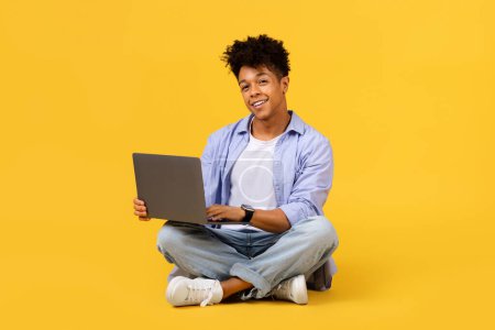 Téléchargez les photos : Joyeux étudiant noir ou pigiste assis jambes croisées sur le sol, travaillant sur un ordinateur portable avec une expression heureuse, sur fond jaune vif - en image libre de droit