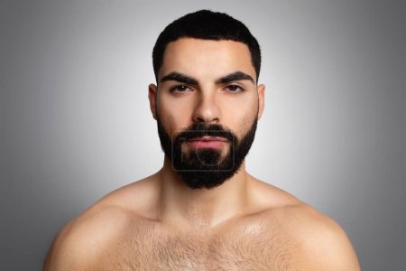 Téléchargez les photos : Portrait rapproché d'un jeune homme arabe athlétique barbu, torse nu et sérieux, posé isolé sur fond de studio gris. Hommes soin de la peau, concept de toilettage de la barbe - en image libre de droit