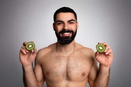 Téléchargez les photos : Boisson vitaminée. Jeune homme du Moyen-Orient à la barbe à moitié nu et positif tenant des kiwis, posant sur fond gris en studio. Alimentation saine, concept de nutrition - en image libre de droit