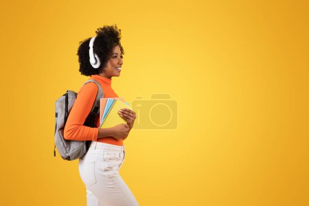 Téléchargez les photos : Vue latérale d'un contenu jeune étudiant afro-américain aux cheveux bouclés portant un casque blanc, un col roulé orange et un sac à dos gris, tenant des carnets sur un fond jaune - en image libre de droit