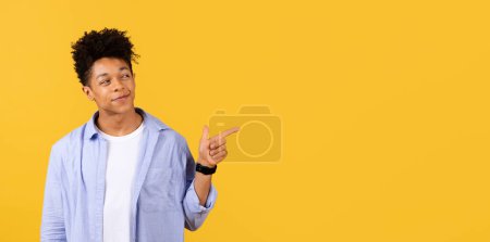 Téléchargez les photos : Homme noir auto-assuré avec les cheveux bouclés pointant vers le côté à l'espace libre avec le sourire connaissant, suggérant ou annonçant quelque chose, sur fond jaune vif, panorama - en image libre de droit
