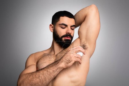 Photo for Middle Eastern Man Applying Deodorant Antiperspirant On Armpit Standing Over Grey Background. Male Hygiene Routine, Sweat And Smell Protection, Skincare Cosmetics - Royalty Free Image