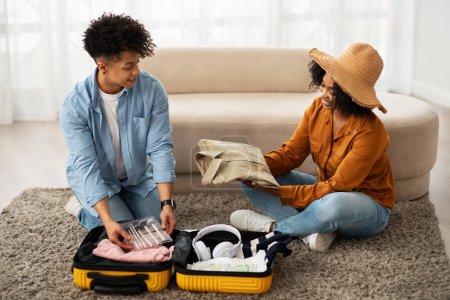 Photo for Engaged millennial African American couple sitting on the floor, joyfully packing clothing into a bright yellow suitcase, preparing for a trip with a sense of adventure and togetherness - Royalty Free Image