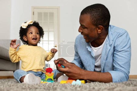 Foto de Feliz joven padre negro jugando junto con su pequeña hija en casa, divirtiéndose con bloques de construcción de juguetes, papá cariñoso disfrutando haciendo actividades de desarrollo con su bebé - Imagen libre de derechos