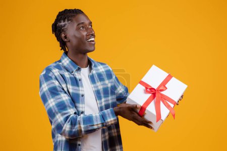Téléchargez les photos : Délicieux jeune homme noir tenant boîte cadeau blanche avec ruban rouge, heureux homme afro-américain regardant vers le côté avec expression joyeuse, portant chemise à carreaux sur fond de studio jaune - en image libre de droit