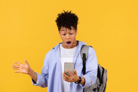 Téléchargez les photos : Étonné étudiant noir masculin exprime son étonnement devant son smartphone, portant un sac à dos, placé sur un fond jaune vif, moment de découverte inattendue - en image libre de droit
