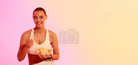 Téléchargez les photos : Concept de mode de vie sain avec une femme rayonnante en tenue de sport, tenant un bol de salade de fruits frais et dégustant une collation nutritive après l'entraînement sur un fond dégradé. - en image libre de droit