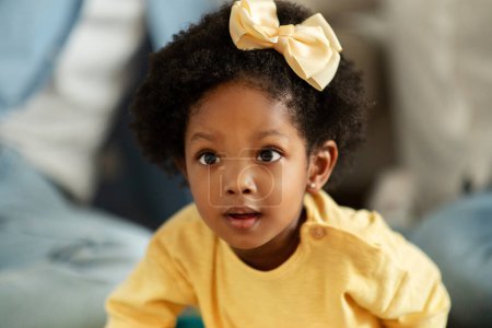 Téléchargez les photos : Gros portrait de mignonne petite fille afro-américaine avec arc de cheveux assis sur le sol à la maison, profiter du temps avec ses parents, fond flou. Concept d'enfance heureuse - en image libre de droit