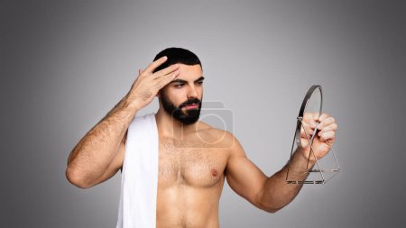 Foto de Caliente guapo barbudo joven de Oriente Medio con toalla en el hombro mirando al espejo de mano después de la ducha, tocando su frente, fondo gris. Primeros signos de envejecimiento de la piel facial - Imagen libre de derechos