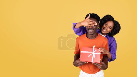 Foto de Una mujer sonriente con una camisa púrpura cubre juguetonamente los ojos de un hombre con una camisa naranja, que sostiene una caja de regalo roja con una cinta blanca, ambas sobre un fondo amarillo brillante - Imagen libre de derechos