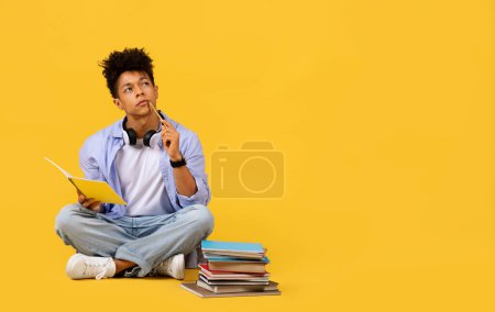 Foto de Estudiante negro pensativo se sienta con las piernas cruzadas con pila de libros, sosteniendo el cuaderno y el lápiz, mirando el espacio libre, sobre el fondo amarillo, bandera - Imagen libre de derechos