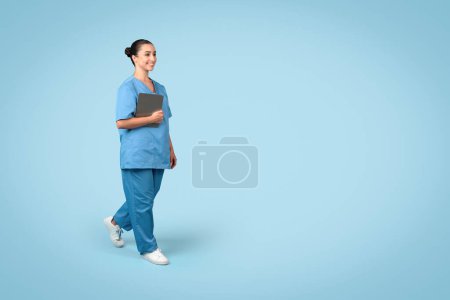 Photo for Cheerful young female nurse in scrubs holding tablet while walking, looking positive and professional, blue background, free space, medical banner - Royalty Free Image