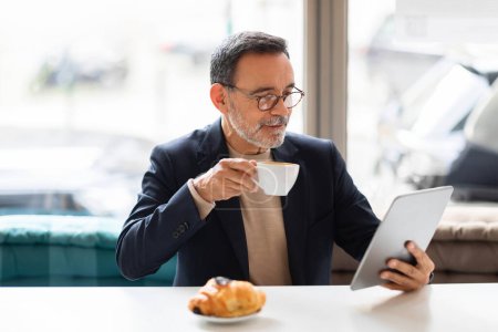 Photo for Sophisticated caucasian mature man in glasses sipping from a white espresso cup while reading on a tablet, with a fresh croissant on the table, in a well-lit cafe setting - Royalty Free Image