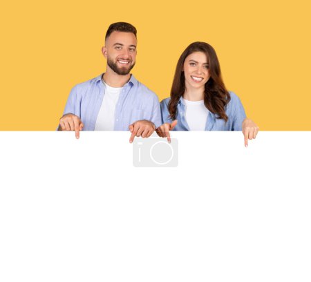 Photo for Happy young man and woman in stylish denim shirts pointing down at large white blank banner with plenty of copy space, against yellow background - Royalty Free Image
