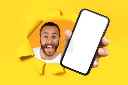 Photo for Enthusiastic bearded man in a white t-shirt joyfully breaks through a yellow paper wall, showing a blank smartphone screen. Recommendation website, app, good news, win and technology - Royalty Free Image