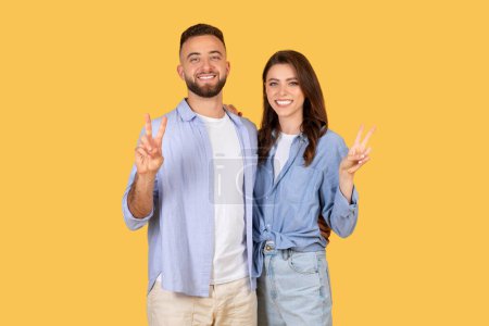 Photo for Radiant european couple posing with peace hand signs, exuding friendliness and positivity, standing closely together against bright yellow background - Royalty Free Image
