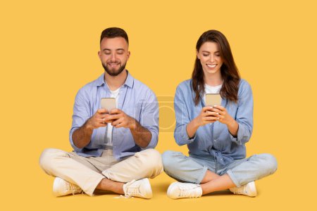 Photo for Cheerful young man and woman sit on floor with legs crossed, each happily using their own smartphones, showcasing modern communication, yellow backdrop - Royalty Free Image