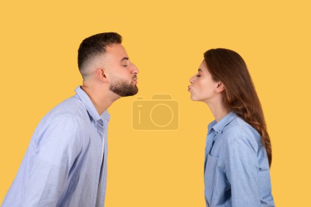 Foto de Pareja joven juguetona de pie cara a cara, acurrucándose para besarse contra un fondo amarillo sólido, con expresiones de afecto y diversión, vista lateral - Imagen libre de derechos