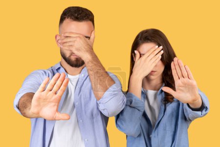 Man and woman covering eyes with one hand while gesturing stop with the other, do not want to see something, standing against yellow background