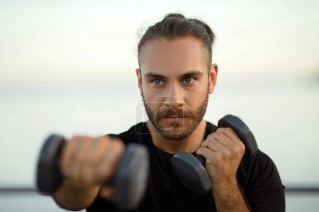 Photo for Determined man in fitwear exercises with dumbbells, embodying active and healthy lifestyle, flexing biceps during morning seaside workout outside. Portrait shot of guy boxing with weights - Royalty Free Image