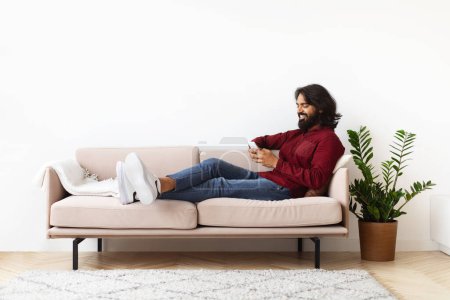 Téléchargez les photos : Cool barbu millénariste indien gars allongé sur le canapé avec smartphone dans ses mains, confortable salon intérieur, textos amis, jeux en ligne, copier l'espace pour la publicité - en image libre de droit