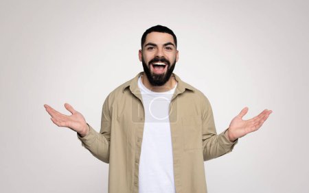 Foto de Un exuberante hombre árabe milenario con barba abre alegremente sus manos, con una camisa beige y una camiseta blanca, expresando un gesto de bienvenida sobre un fondo suave - Imagen libre de derechos