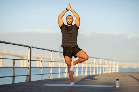 Photo for Fit young man in black sportswear strikes the Tree Pose practicing yoga on boardwalk by the sea, exercise promotes wellbeing and healthy lifestyle, showcasing strength and balance. Full length shot - Royalty Free Image