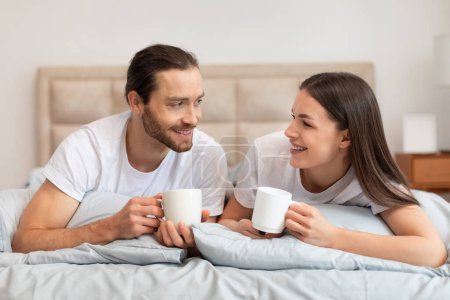 Téléchargez les photos : Couple détendu et heureux en t-shirts blancs profitant de leur café du matin au lit, échangeant des sourires et profitant de l'autre compagnie dans une chambre sereine - en image libre de droit