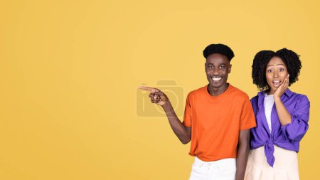 Téléchargez les photos : Un jeune homme animé en chemise orange pointe sur le côté tandis qu'une jeune femme surprise en chemisier violet réagit avec excitation, à la fois sur un fond jaune vif - en image libre de droit