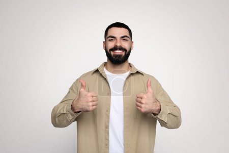 Téléchargez les photos : Optimiste homme arabe barbu millénaire en chemise beige et t-shirt blanc donnant deux pouces vers le haut avec un large sourire, indiquant le succès ou l'approbation sur un fond clair, studio - en image libre de droit