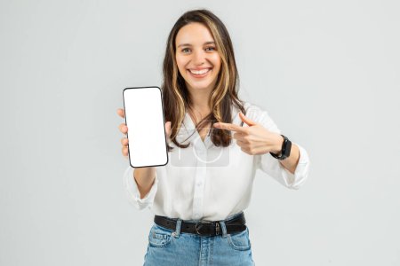 Photo for Cheerful woman in a white blouse and blue jeans showing a smartphone with a blank screen, pointing at it with her finger, wearing a smartwatch, on a light grey background - Royalty Free Image
