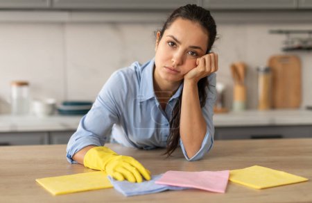 Mujer joven cansada con guantes amarillos apoya su mejilla en su mano mientras limpia su casa, su expresión transmite mezcla de fatiga y aburrimiento en la cocina ordenada