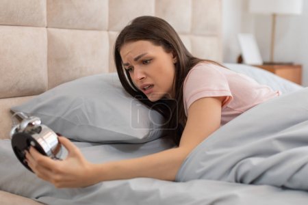 Mujer joven angustiada en la cama, frunciendo el ceño ante el ruidoso anillo del reloj despertador clásico, capturando la lucha matutina de despertarse temprano
