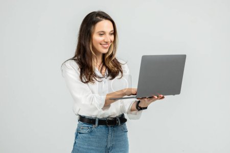 Téléchargez les photos : Jeune femme engagée dans un chemisier blanc et un jean bleu centré sur un ordinateur portable gris qu'elle tient, avec un sourire subtil et une montre intelligente au poignet, debout sur une toile de fond grise - en image libre de droit