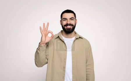 Téléchargez les photos : Jeune homme arabe confiant et souriant avec une barbe qui fait un excellent signe avec sa main, portant une chemise beige sur un t-shirt blanc, debout sur un fond uni, studio - en image libre de droit