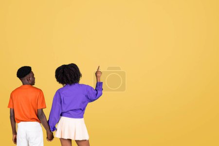 Téléchargez les photos : Vue arrière d'un jeune couple connecté, la femme en chemisier violet pointant vers le haut tout en tenant la main d'un homme en chemise orange, sur un fond jaune minimaliste - en image libre de droit