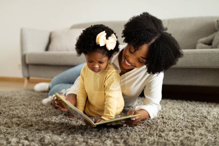 Téléchargez les photos : Temps de jeu. Joyeux amour afro-américain mère lecture livre avec sa petite fille, fille pointant vers des images colorées, passer du temps libre ensemble à l'intérieur de la maison, espace de copie - en image libre de droit