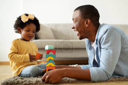 Téléchargez les photos : Temps de jeu. Joyeux aimant jeune père noir jouant des jouets colorés avec sa petite fille, tour de bâtiment, fille en mouvement cubes, passer du temps libre ensemble à l'intérieur de la maison, espace de copie - en image libre de droit