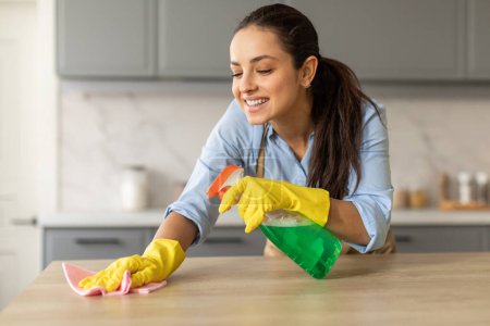 Foto de Joven alegre con guantes amarillos y camisa azul limpia la encimera de la cocina con paño rosa y botella de spray verde, disfrutando de las tareas domésticas - Imagen libre de derechos