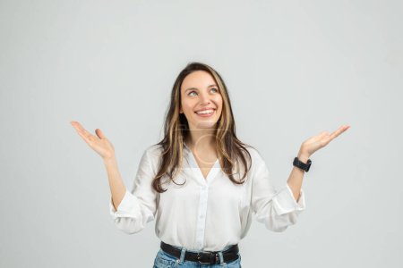 Photo for An upbeat smiling european young woman in a white blouse and blue jeans is raising her hands, palms up, in a gesture of uncertainty or weighing options, with a smile on her face - Royalty Free Image