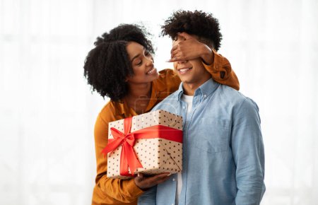 Photo for An affectionate African American millennial woman surprises her partner with a covered eyes gesture and a large heart-patterned gift box adorned with a red ribbon, conveying love and happiness - Royalty Free Image