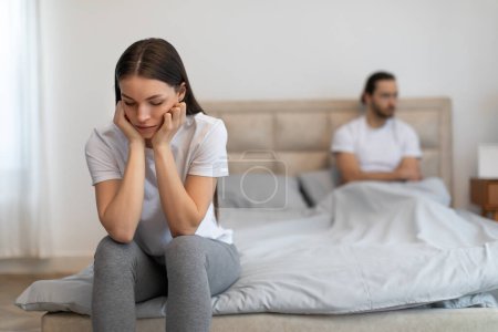 Gestresste junge Frau sitzt mit dem Kopf in den Händen auf dem Bett, während sich der Mann auf dem Hintergrund ausruht und den Moment der Besorgnis oder Uneinigkeit einfängt, Schlafzimmereinrichtung