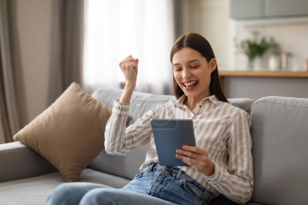 Photo for Delighted young woman sitting on couch, triumphantly raises her fist while looking at digital tablet, expressing joy and achievement, reading great news - Royalty Free Image