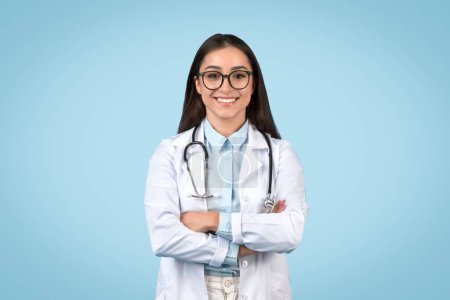 Foto de Alegre joven doctora con gafas, usando estetoscopio y bata blanca de laboratorio, brazos cruzados, sonriendo a la cámara sobre fondo azul - Imagen libre de derechos