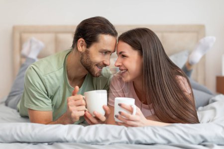 Photo for Happy, relaxed couple lying in bed with white mugs, engaging in joyful, intimate conversation while smiling, touching forehead and making eye contact - Royalty Free Image