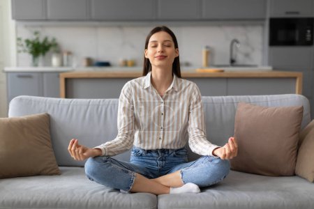 Photo for Serene young woman in casual attire enjoying moment of relaxation while practicing meditation seated on comfortable sofa in well-lit living room - Royalty Free Image
