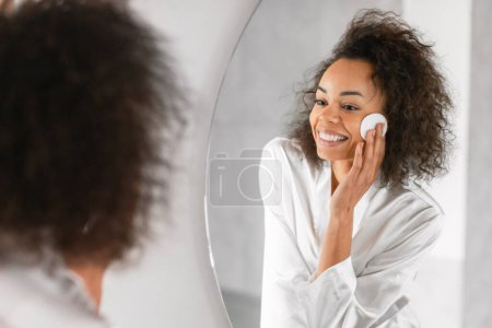 Photo for African American woman takes care of her skin with cotton pad and lotion, moisturizing and cleansing face near mirror at domestic bathroom indoor, revealing radiant complexion. Selective focus - Royalty Free Image